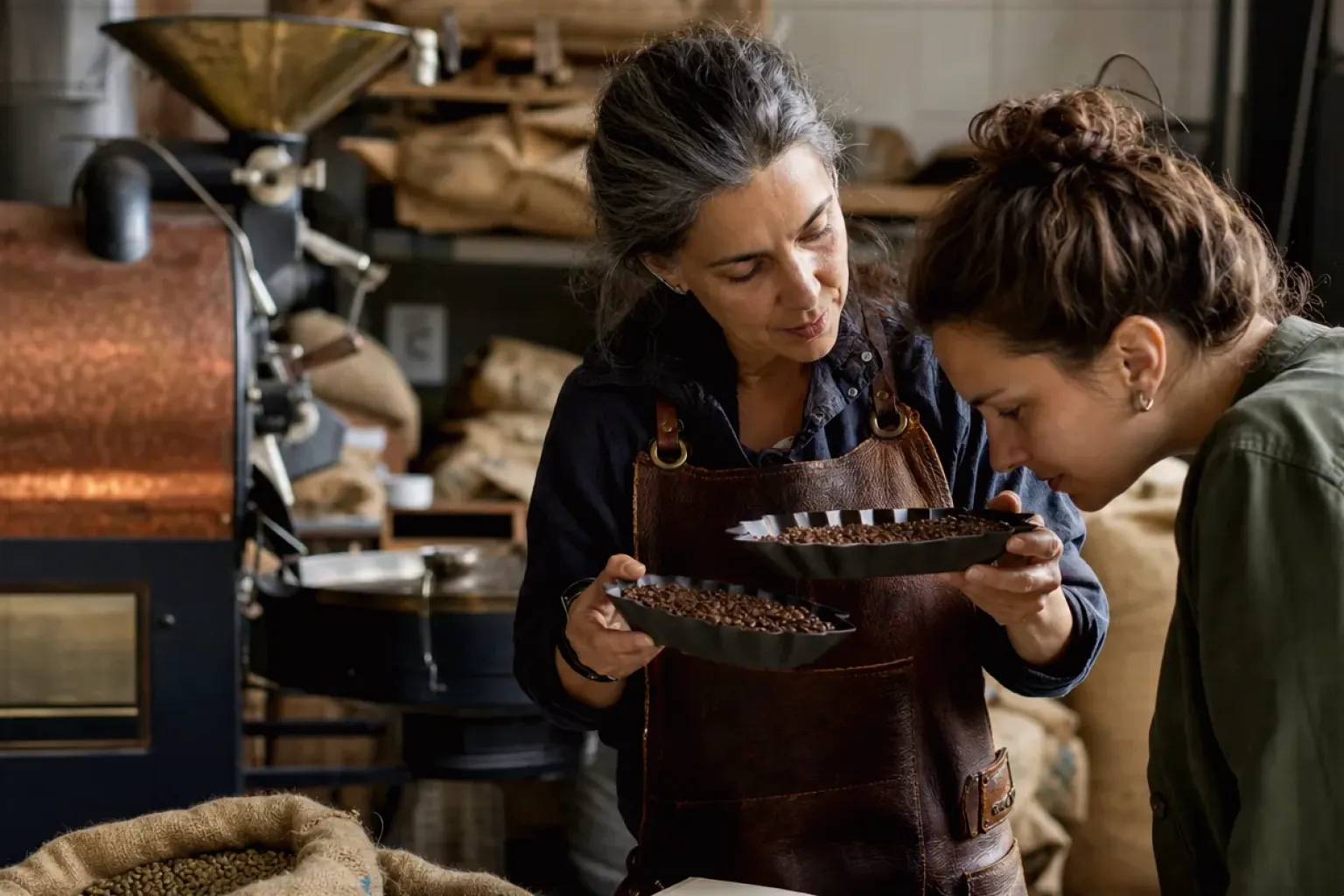 Eine Kaffeerösterin lässt eine Kundin in einer kleinen Rösterei verschiedene frisch geröstete Bohnen riechen – gezeigt für Handfilter im Themenvergleich.